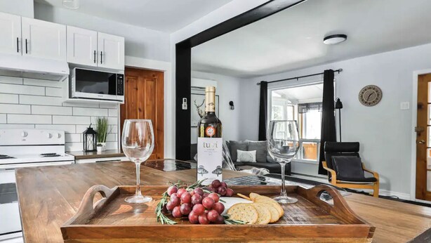 A modern kitchen with white cabinets, a wooden countertop, and a wine bottle on a tray with grapes and crackers. - Chalet with waterfront, Spa and magnificent view of a regional park (Chertsey)