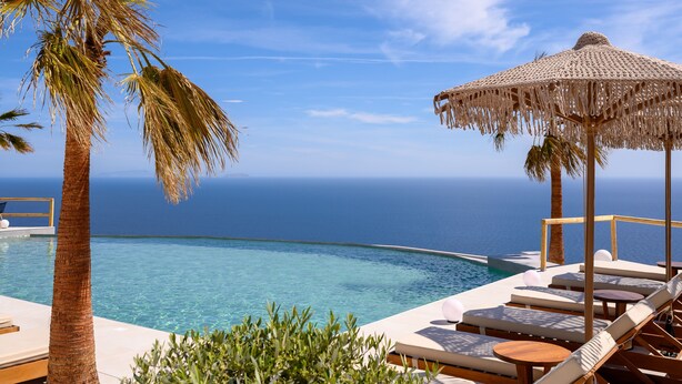 Un espace aménagé au bord de la piscine, avec des chaises longues, un parasol en chaume et une vue sur l’océan.