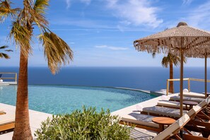 Un área junto a la piscina con sillones de descanso, una sombrilla de paja y vista al océano.