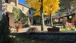 A courtyard with stone buildings, a tree with yellow leaves, and potted plants. - Typical stone cottage with large garden and private parking space (Passignano sul Trasimeno)