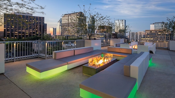 Une terrasse sur le toit, dotée de mobilier contemporain et agrémentée d’un foyer central, le tout surplombant un panorama urbain au crépuscule.