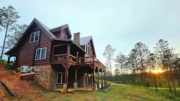 A large, two-story log cabin with a stone chimney, a porch with wooden railings, and a view of the sunset.