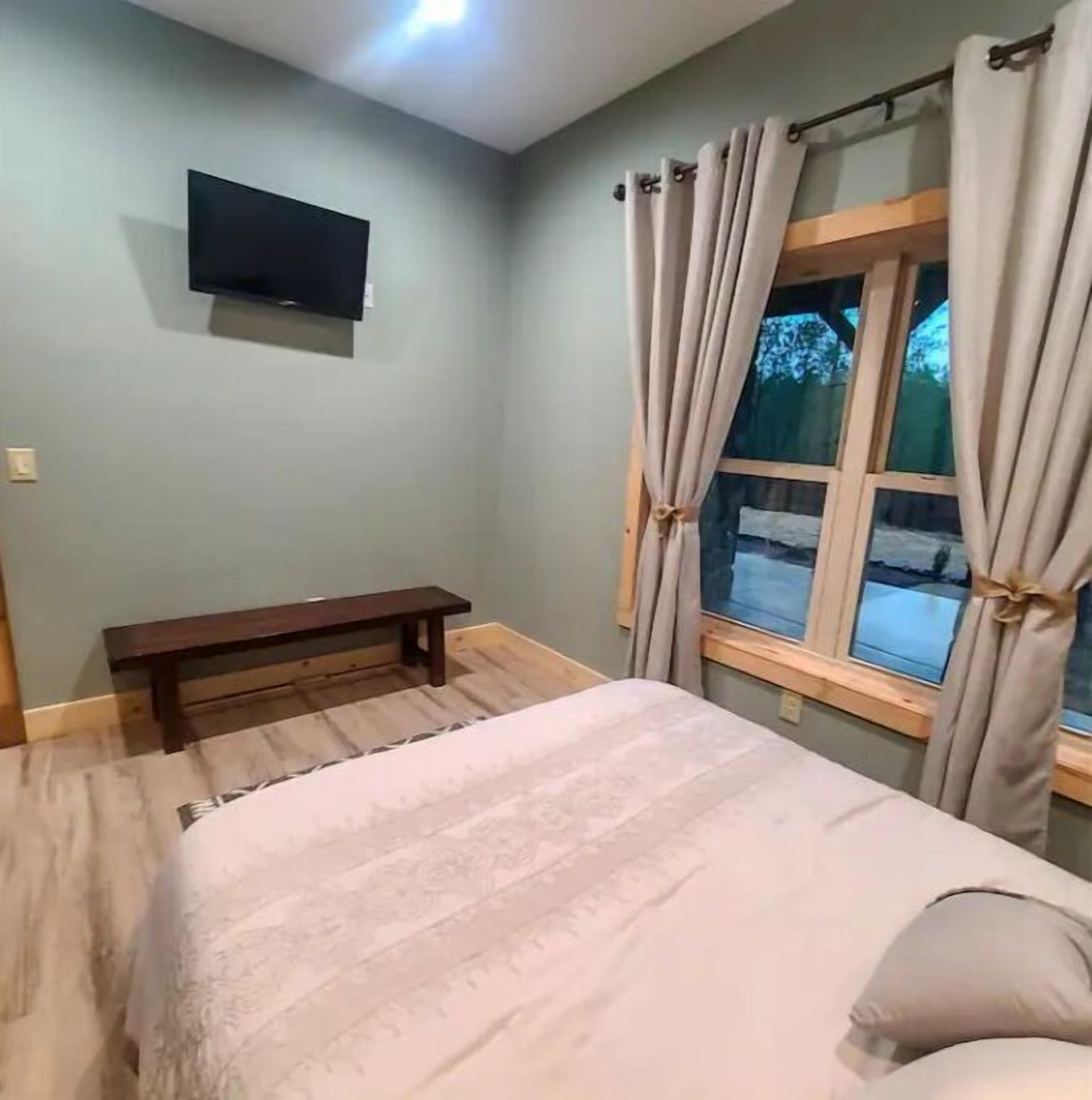 A bedroom with a bed, a bench, a TV mounted on the wall, and a window with curtains.