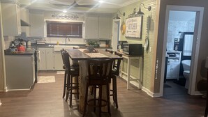 A spacious kitchen with a central island, wooden dining table, and chairs. There is a visible refrigerator, stove, and sink area. - The Pink House - Charming, fully renovated, quiet getaway (Wellfleet)