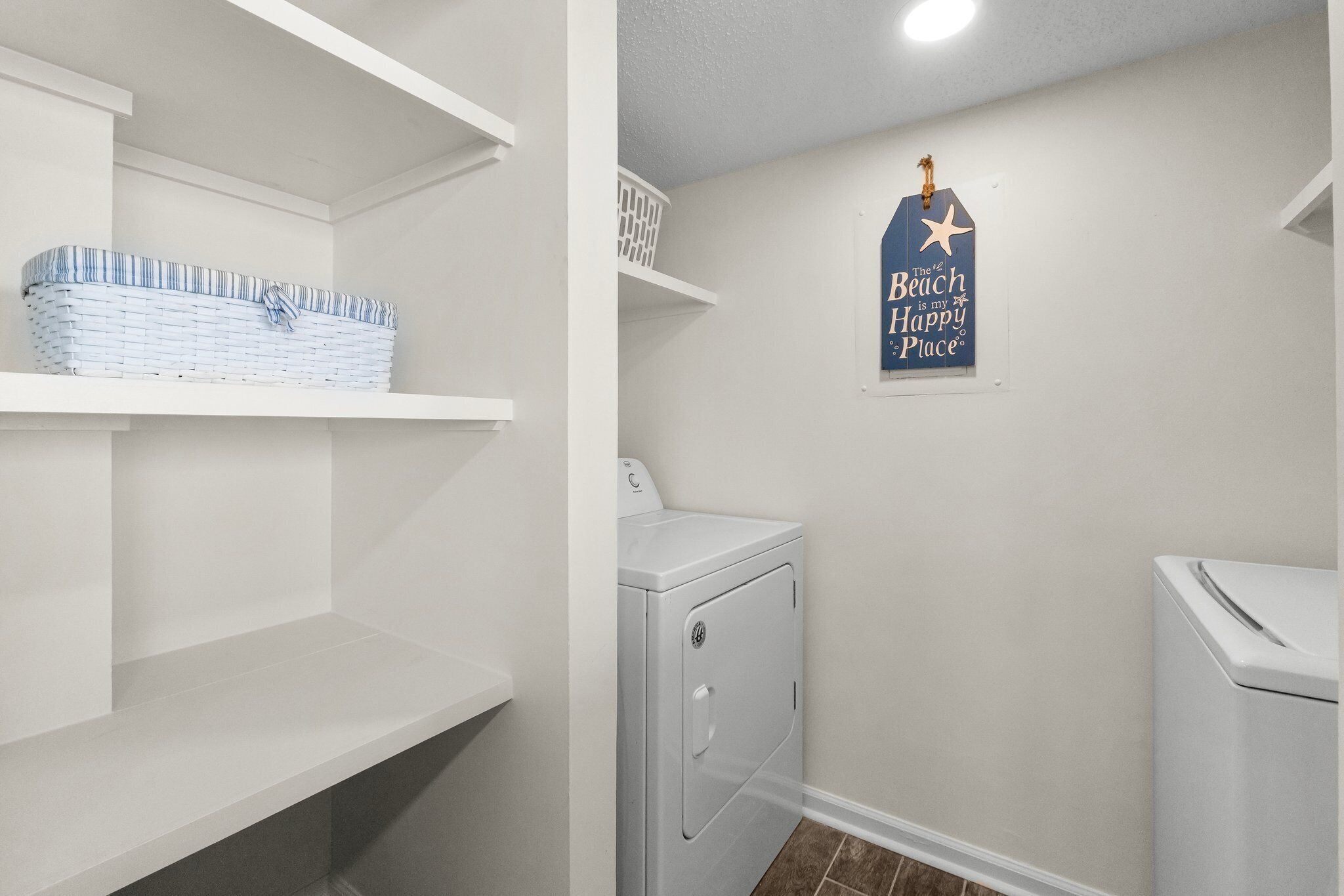 A laundry room with a stacked washer and dryer, a basket on a shelf, and a wall sign that reads "The Beach is my Happy Place."