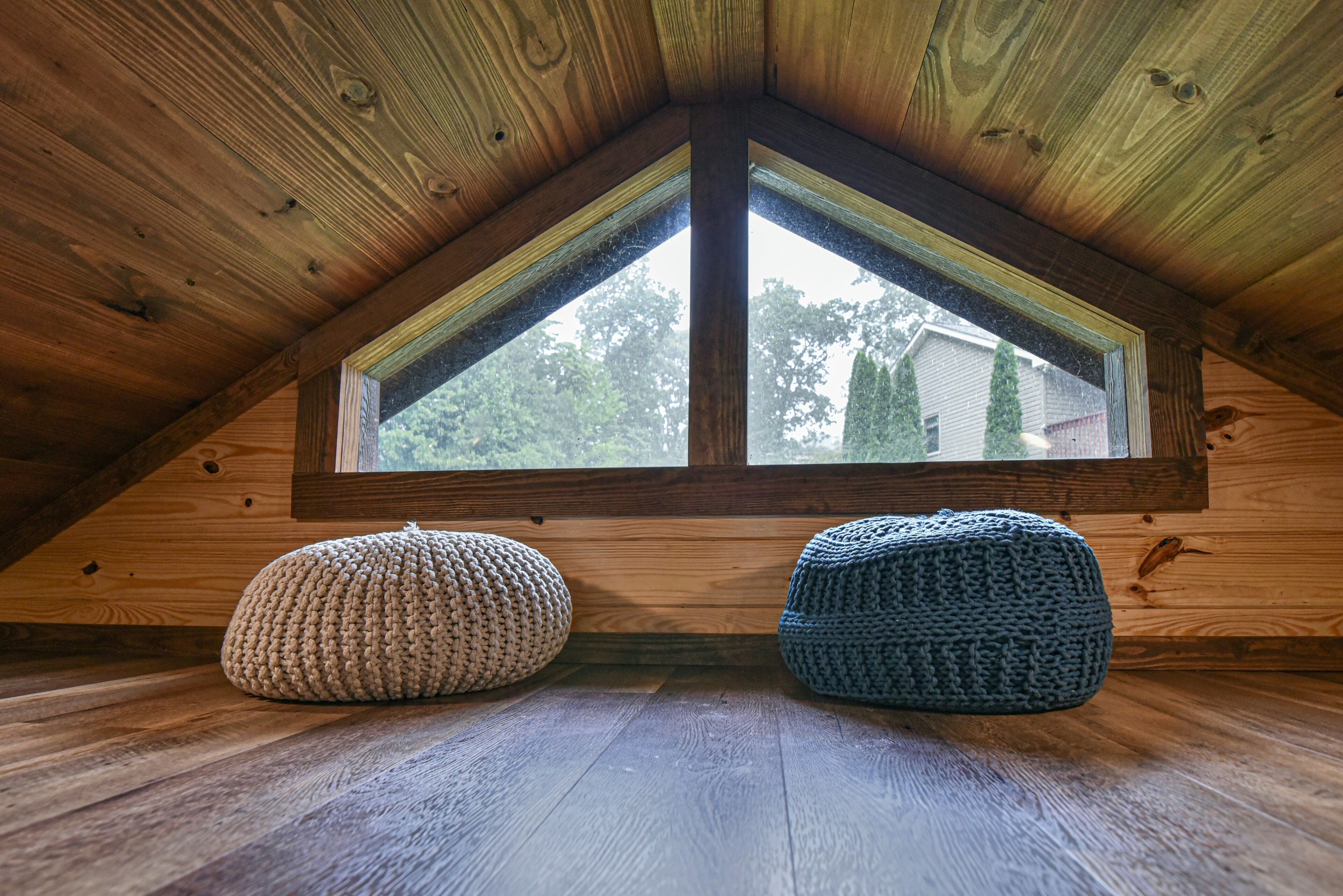 Une pièce mansardée en bois, avec un plafond en pente et une grande fenêtre. Deux poufs tricotés sont posés sur le sol.