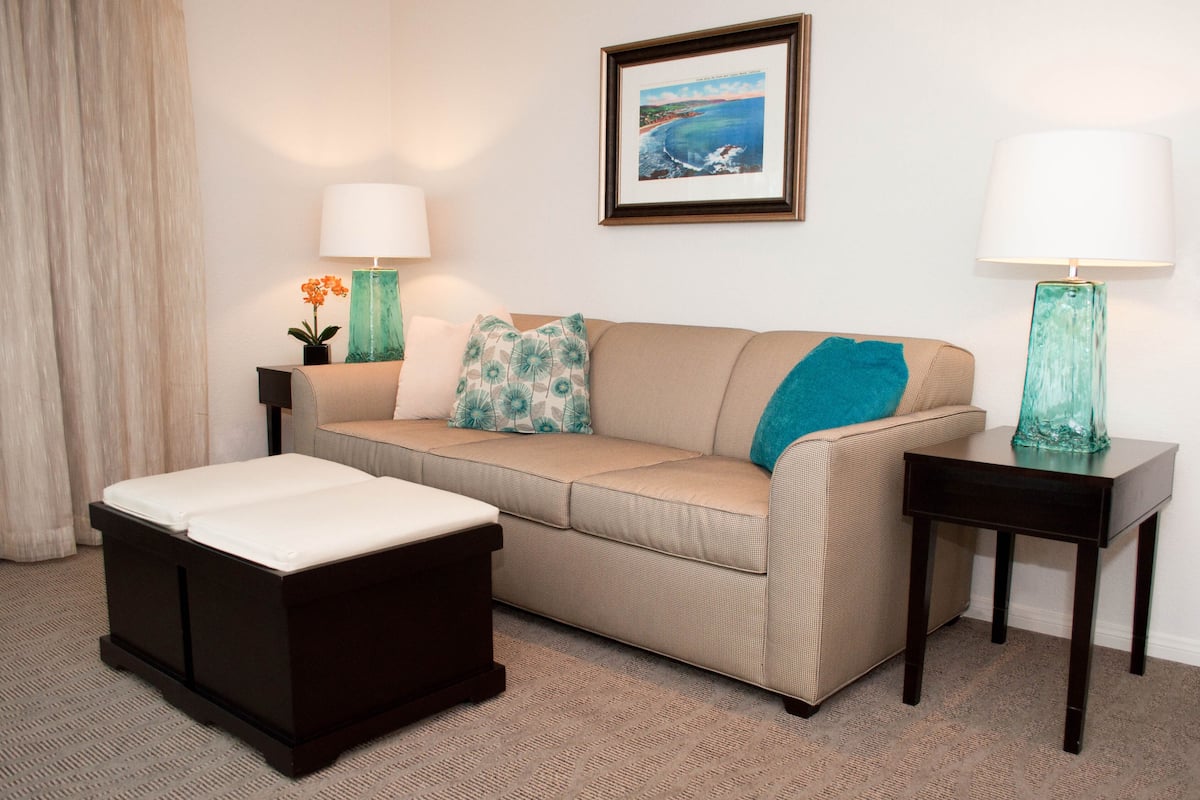 A living room with a beige sofa, a dark ottoman, two side tables with lamps, a framed picture on the wall, and a potted plant.