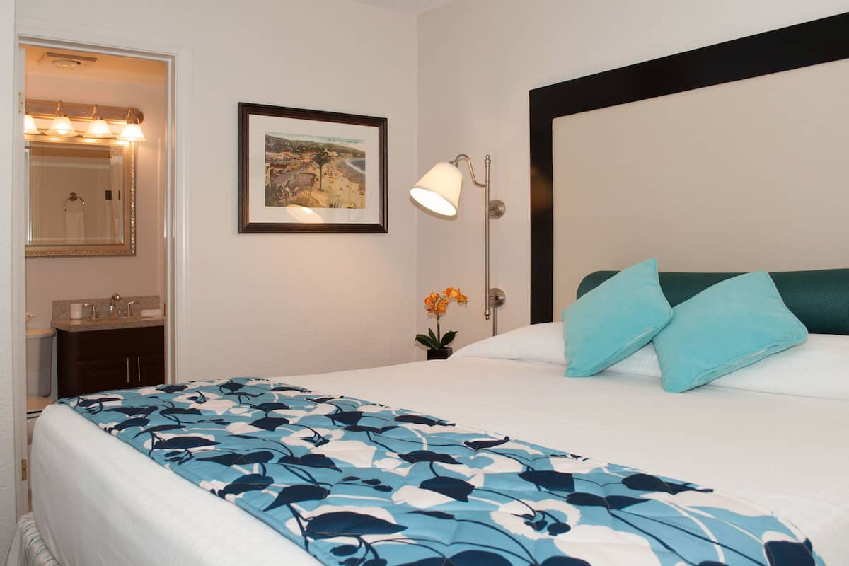 A hotel room with a bed, a nightstand with a lamp, a framed picture on the wall, and a bathroom visible through an open door.