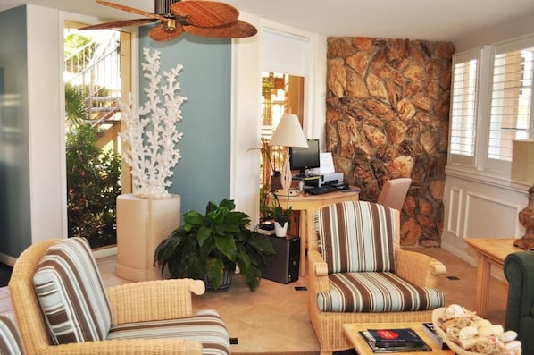 A cozy living room with wicker furniture, a stone wall, and a view of a balcony with greenery. - Laguna Shores Studio Suites (Laguna Beach)