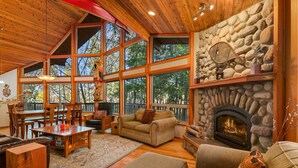 A cozy living room with a stone fireplace, wooden furniture, and large windows offering a view of the outdoors. - Enchanting Lindal Waterfront Cabin (Shelton)