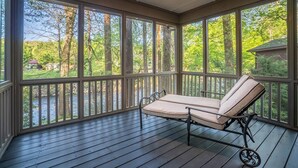 A covered porch with a cushioned bench, large windows, and a view of a river and trees. - Luxury Riverfront Retreat | Hot Tub | Walk to Town (Helen)