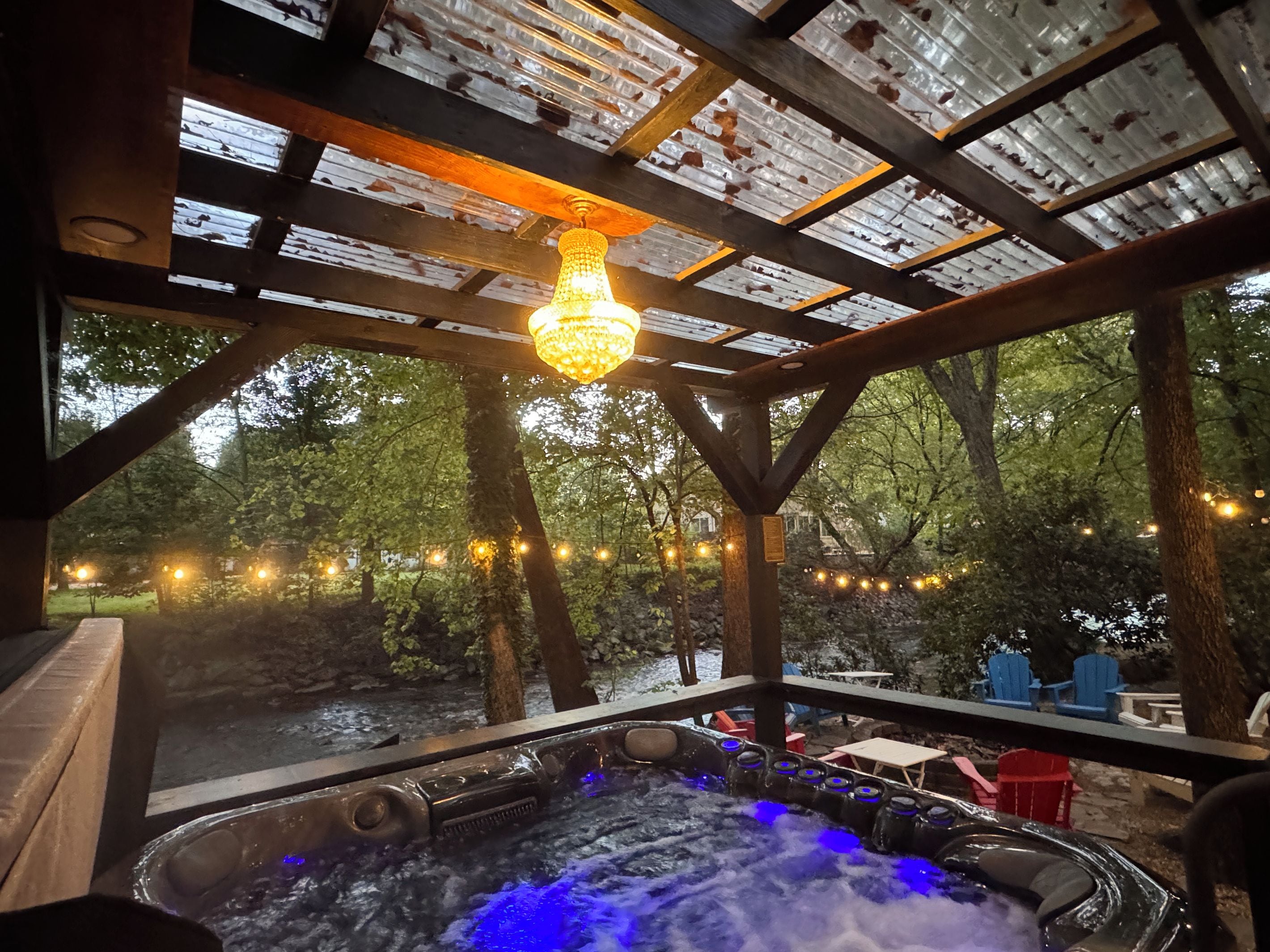 An outdoor hot tub area with a pergola structure, string lights, and a view of the surrounding forest.