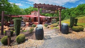 An outdoor seating area with a pergola, tables, and chairs, surrounded by neatly trimmed bushes and a gravel path. - Luxury Riverfront Retreat | Hot Tub | Walk to Town (Helen)