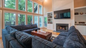 A spacious living room with a large sectional sofa, a fireplace, and a TV mounted on the wall. - Luxury Riverfront Retreat | Hot Tub | Walk to Town (Helen)