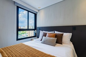 A modern hotel room with a large bed, a view of the city through a window, and a dark headboard.
