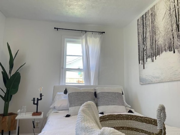 Ein Schlafzimmer mit einem Bett, einem Fenster mit Vorhängen, einem Bild an der Wand und einer Pflanze.