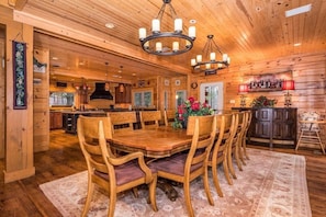 Un intérieur en bois avec une table de salle à manger ronde entourée de chaises, un lustre et un coin cuisine à l’arrière-plan.