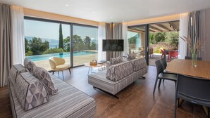 A spacious living area with a large sofa, a television, and a dining table. There is a view of a swimming pool and outdoor seating. - BITTER ALMOND TREES VILLAS - Stylish villa, sea views 1000m from Nikiana & Beach (Lefkada)