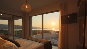 A bedroom with a large bed, a television, and a view of the sunset through floor-to-ceiling windows.
