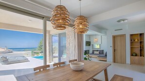 A modern dining area with a wooden table and chairs, a bowl on the table, and a view of the sea through large windows.