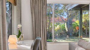 Una habitación con un ventanal grande, una mesa con una lámpara y flores, y vista a un jardín y una casa.