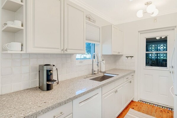 A modern kitchen with white cabinets, a granite countertop, a coffee maker, and a window with blinds. - SoSoPalmBeach-Tropical Bungalow (West Palm Beach)