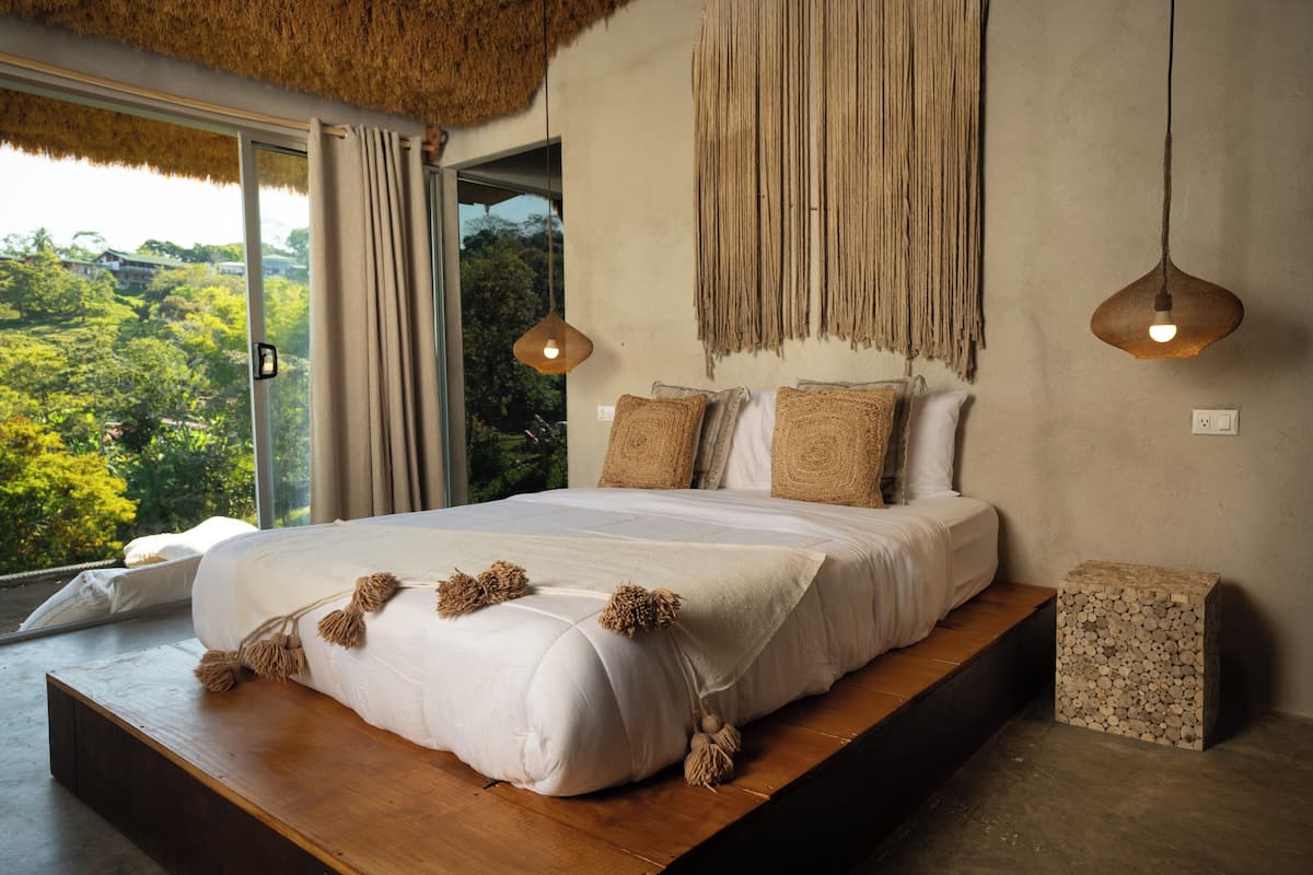 A modern bedroom with a large bed, a view of greenery outside, and a unique textured wall hanging.