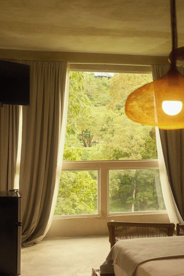 A bedroom with a large window, curtains, a bed, a bedside table, and a lamp.