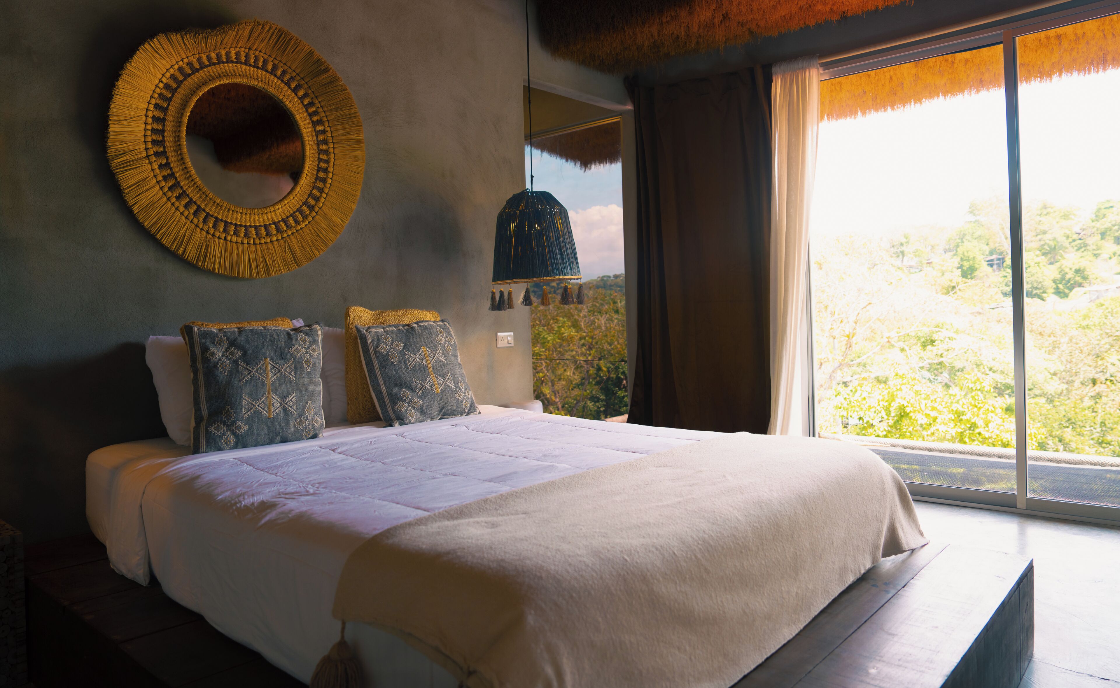 Un letto con copripiumino bianco, due cuscini con rivestimenti decorati, un ornamento circolare a parete e vista su del verde attraverso una finestra.