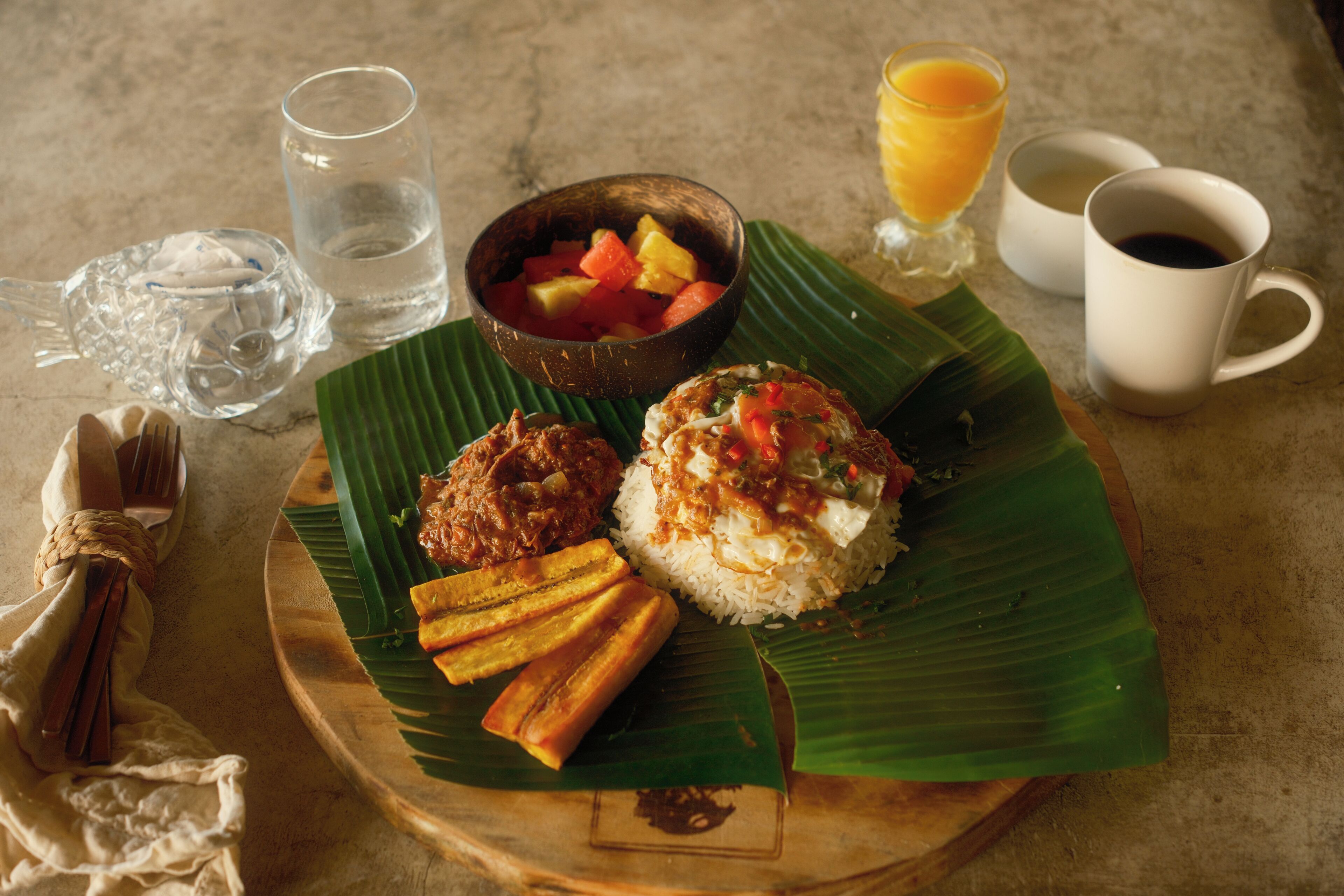 A meal served on a banana leaf with a fork, a glass of water, a bowl of fruit, and two cups of coffee.