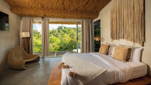 Een slaapkamer met een groot bed, een geweven stoel en uitzicht op groen buiten het raam.