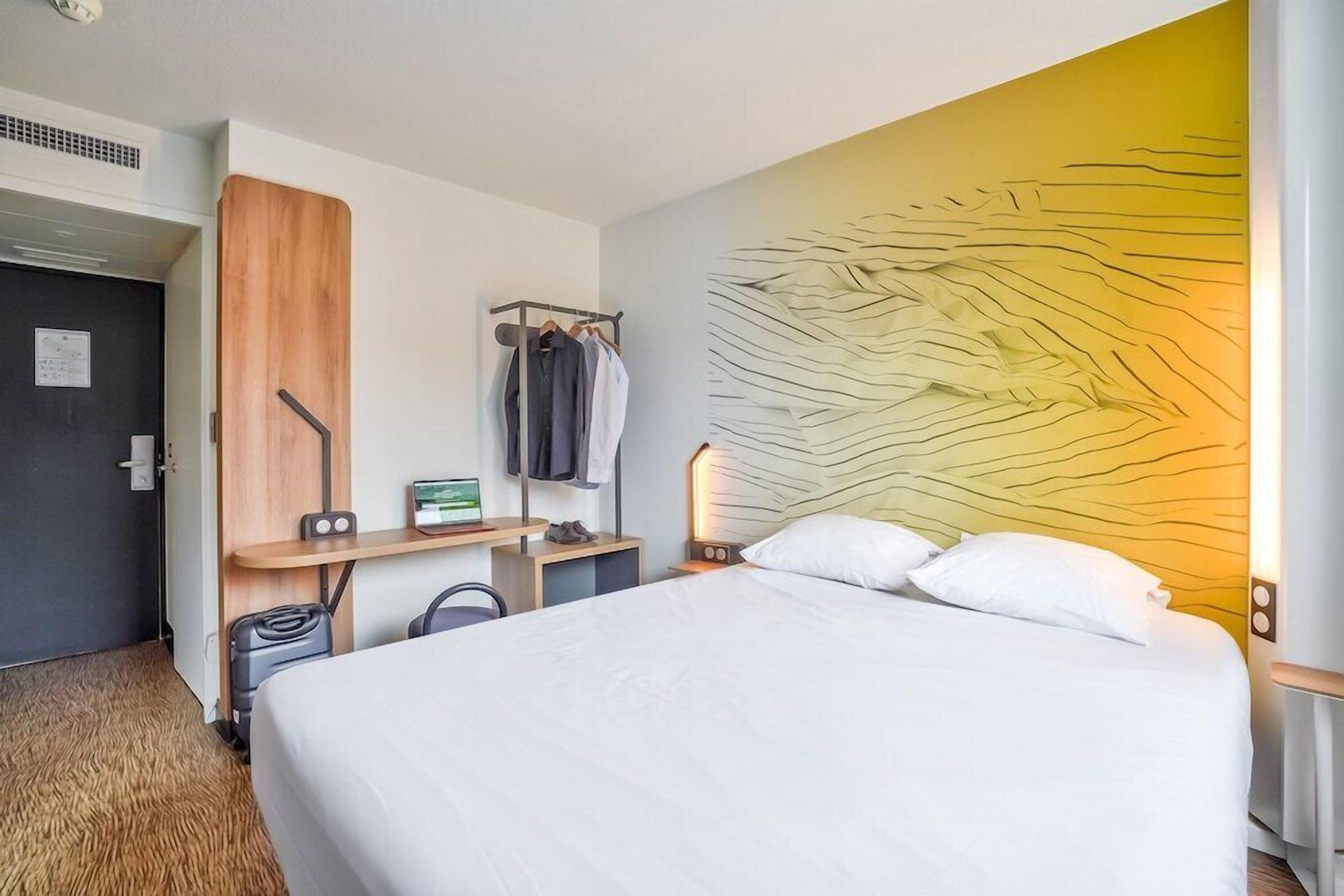 Quarto de hotel com uma cama, uma escrivaninha, um cabideiro, uma luminária na parede e uma porta.