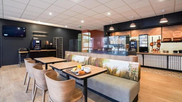 A modern cafe with wooden tables and chairs, a counter with a coffee machine, and a TV mounted on the wall. - B&B Hotel Paris Rosny-sous-bois (Rosny-sous-Bois)