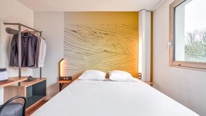 A hotel room with a bed, a desk with a chair, a coat rack with clothes, and a window. - B&B Hotel Paris Rosny-sous-bois (Rosny-sous-Bois)