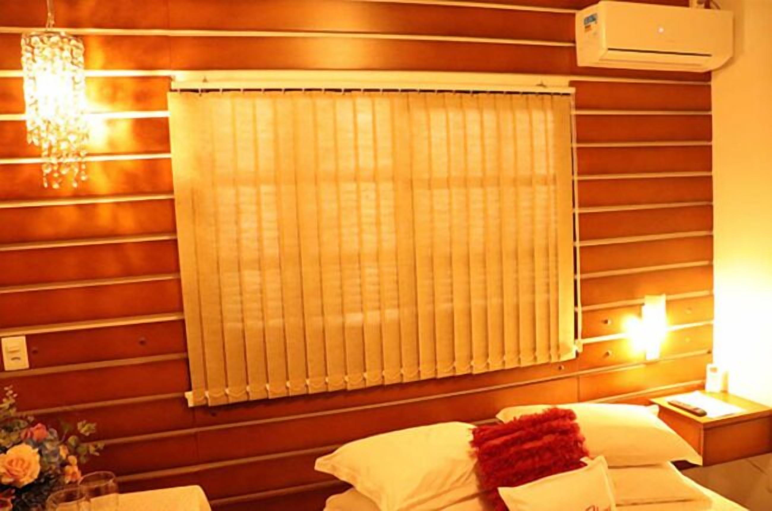 A hotel room with a wooden wall panel, a window with blinds, a bedside table with a lamp, and a small nightstand.