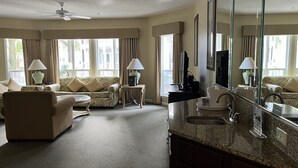 Uma sala de estar ampla com sofá, poltrona e mesa de centro. Há uma televisão, uma mesinha de cabeceira com abajur e um espelho grande. O chão está coberto por um tapete e as janelas são grandes, com cortinas.