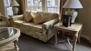 Uma sala de estar bem iluminada com um sofá estampado com motivos florais, duas mesas laterais com abajures e uma mesa de centro com tampo de vidro.
