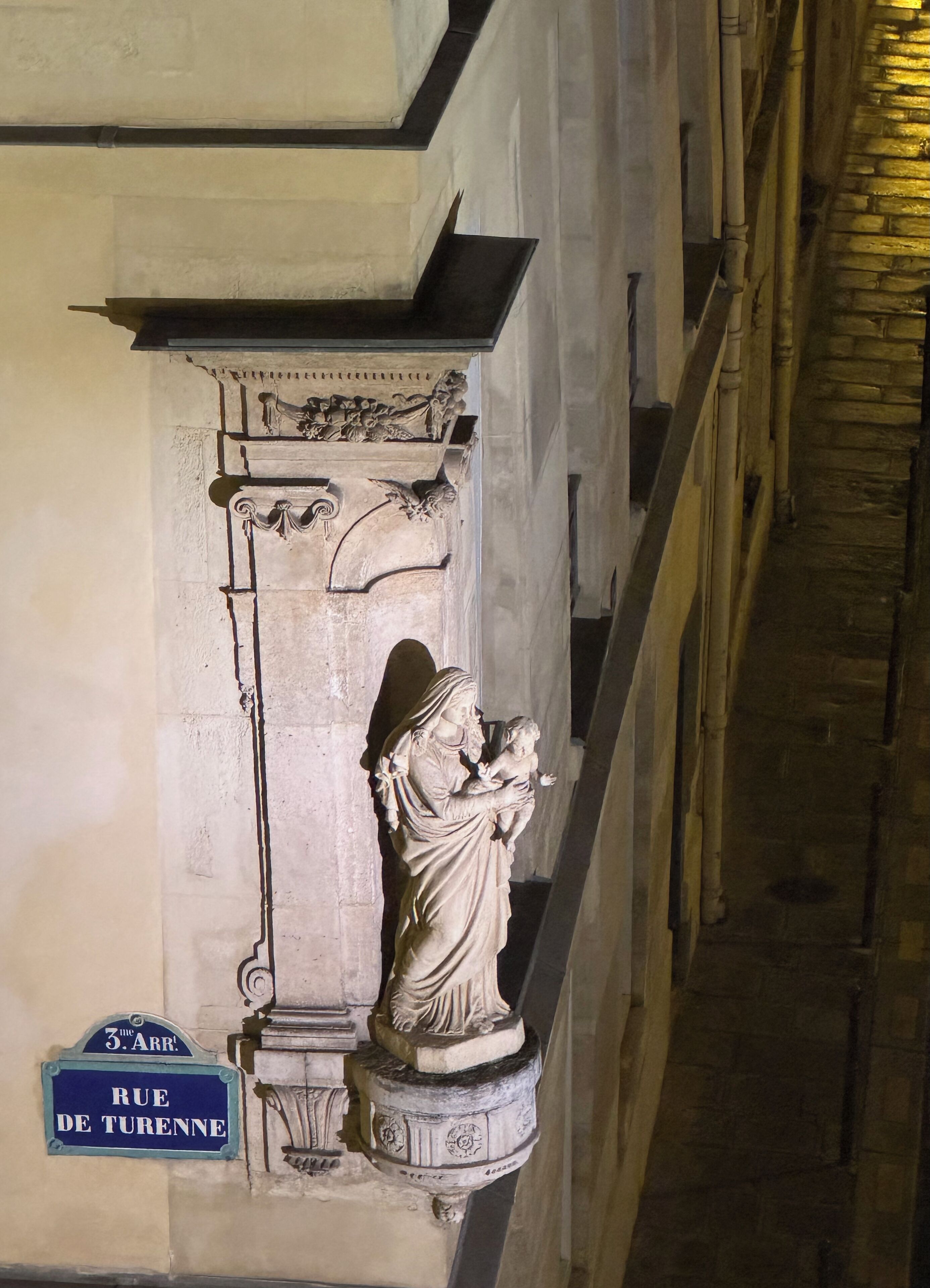 Une statue représentant une femme tenant un enfant, avec une plaque de rue indiquant « RUE DE TURENNE » en dessous.