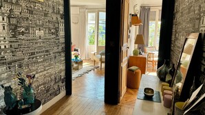 A room with a patterned wall, wooden flooring, a sofa, a chair, and a table with a lamp. - CENTRE MARAIS (picasso place des vosges) .A/C, WIFI, ELEVATOR, VUE, BALCONY ! (paris)