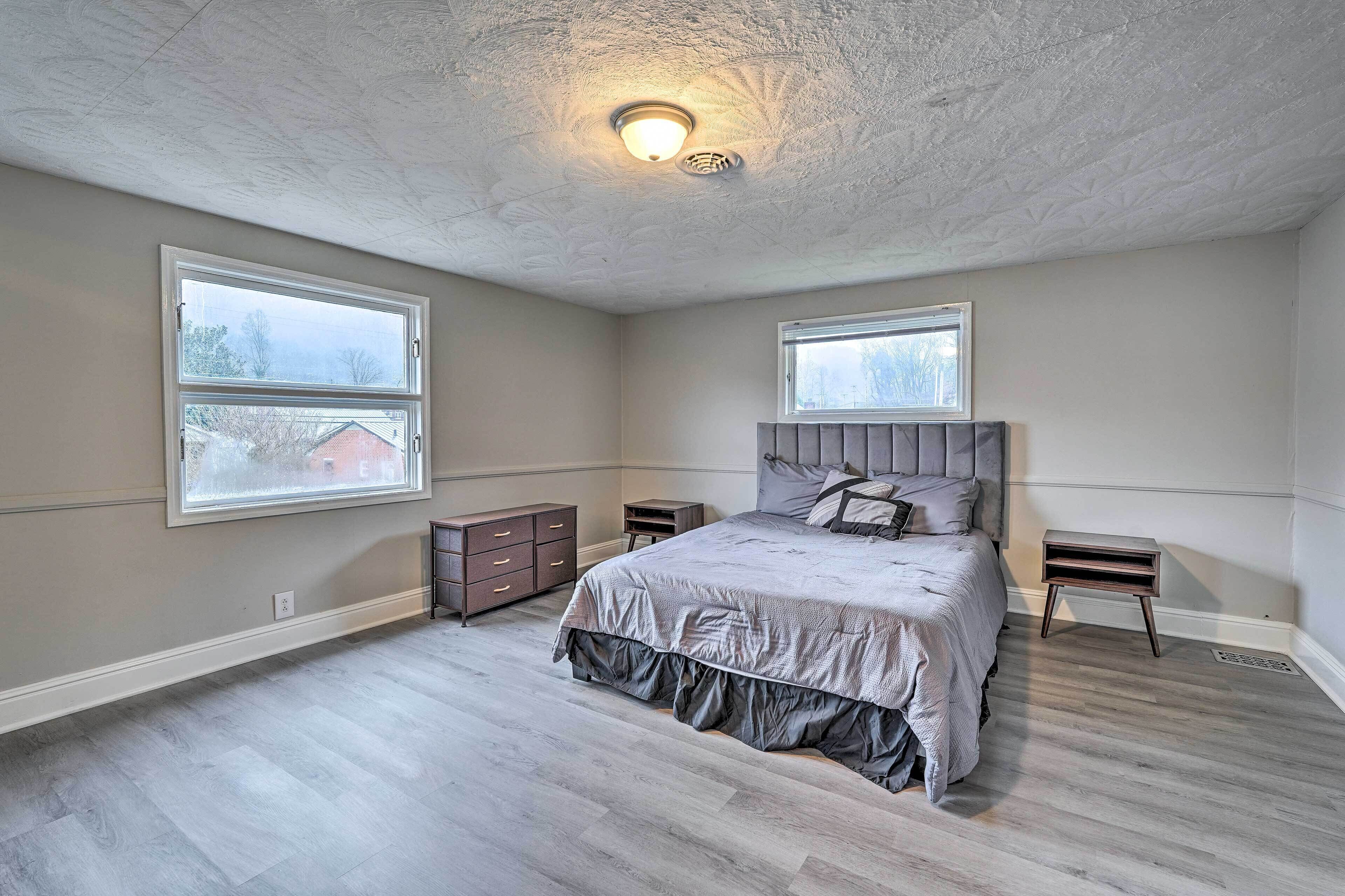 Une chambre à coucher avec un grand lit, deux tables de chevet, un luminaire au plafond et deux fenêtres.