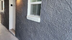 Facciata di un edificio con parete grigia e strutturata, una porta bianca e una finestra con persiane.