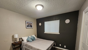Una camera da letto con un letto, un comodino, un quadro appeso al muro, una finestra con persiane e una lampada a soffitto.