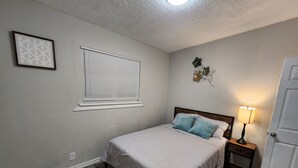 A bedroom with a bed, a window with blinds, a nightstand with a lamp, and a door.
