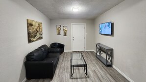 A modern living room with a black leather sofa, a glass coffee table, and a flat-screen TV mounted on the wall.