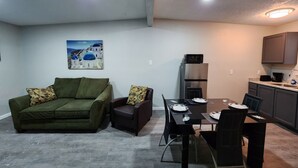 A compact living space with a green sofa, a small table with chairs, a kitchenette with a refrigerator and microwave, and a painting of a coastal scene. - Luxury Apartment Suite 8 (El Paso)