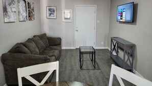 A living room with a grey couch, a small table, a door, and a TV mounted on the wall.
