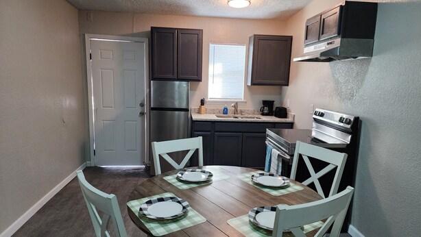 A compact kitchen with dark cabinets, a stainless steel refrigerator, a stove, and a dining table set with plates and placemats.