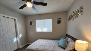 A bedroom with a bed, a ceiling fan, a window with blinds, and a wall-mounted air vent.