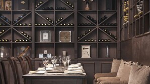 A dining area with a table set for four, surrounded by dark wooden chairs. A sofa with cushions is positioned to the right. The walls are lined with shelves holding wine bottles and framed items. - Apartment B25, Bansko, Royalpark (Bansko)