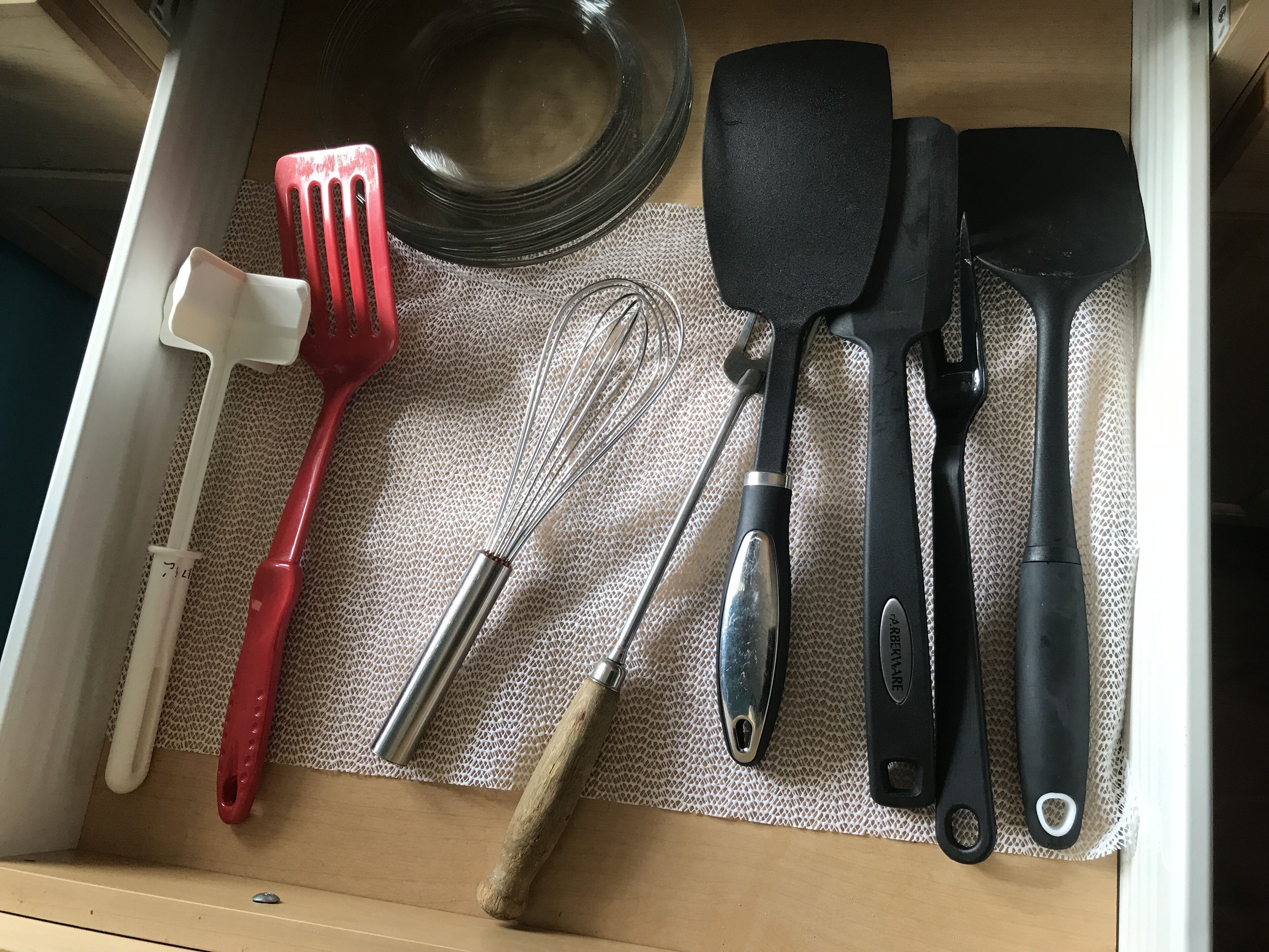 En kökslåda med olika köksredskap, inklusive en röd stekspade, en visp, en svart stekspade och en svart stekspade med ett hål.